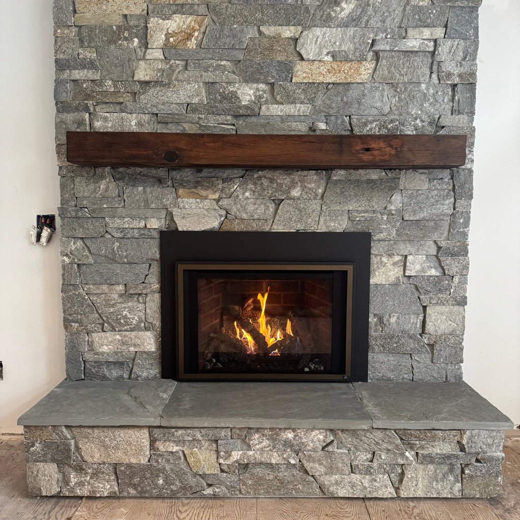 A stone fireplace insert with a black frame showing a glowing fire, set against a gray stone veneer wall with a dark wooden beam on the left.