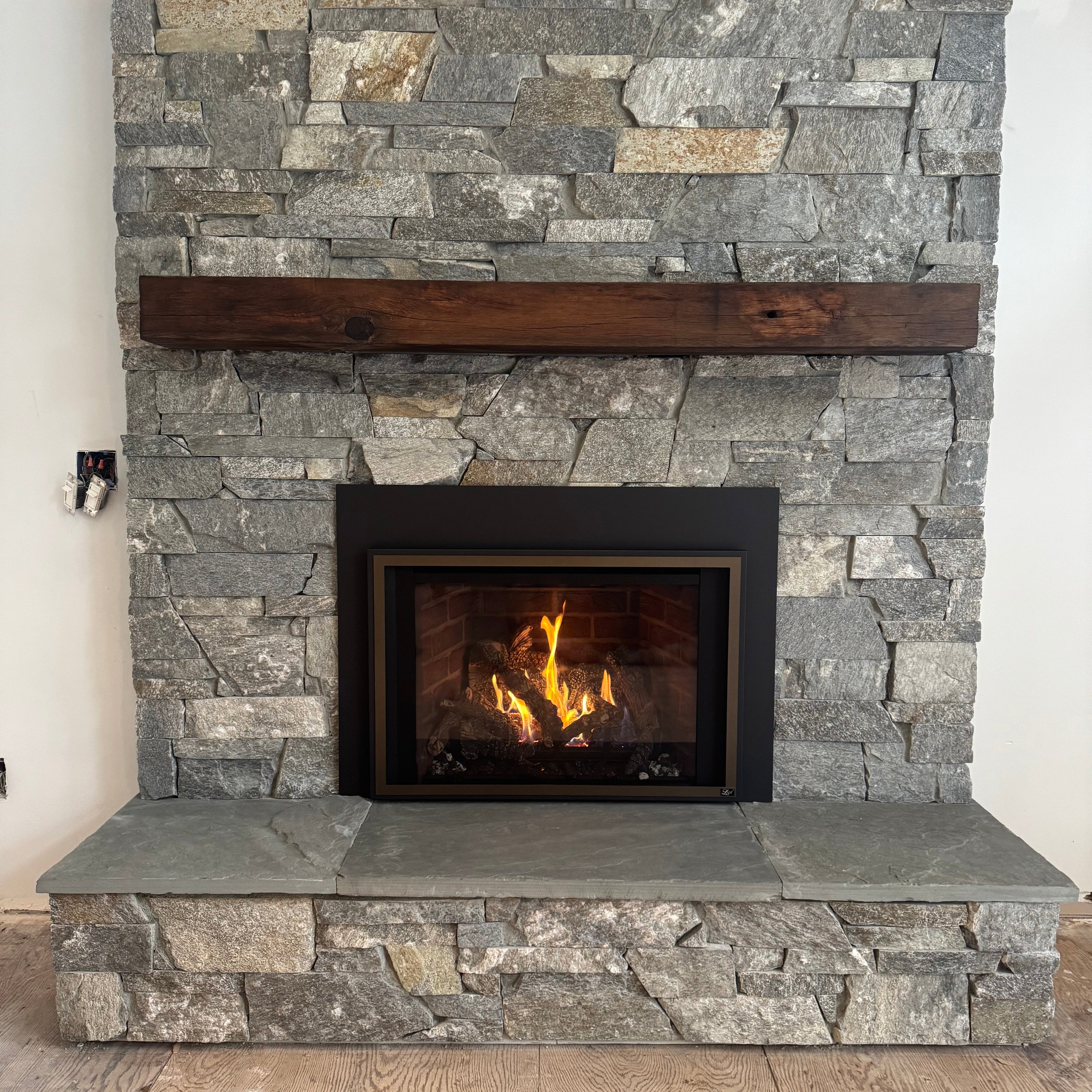 A stone fireplace insert with a black frame showing a glowing fire, set against a gray stone veneer wall with a dark wooden beam on the left.