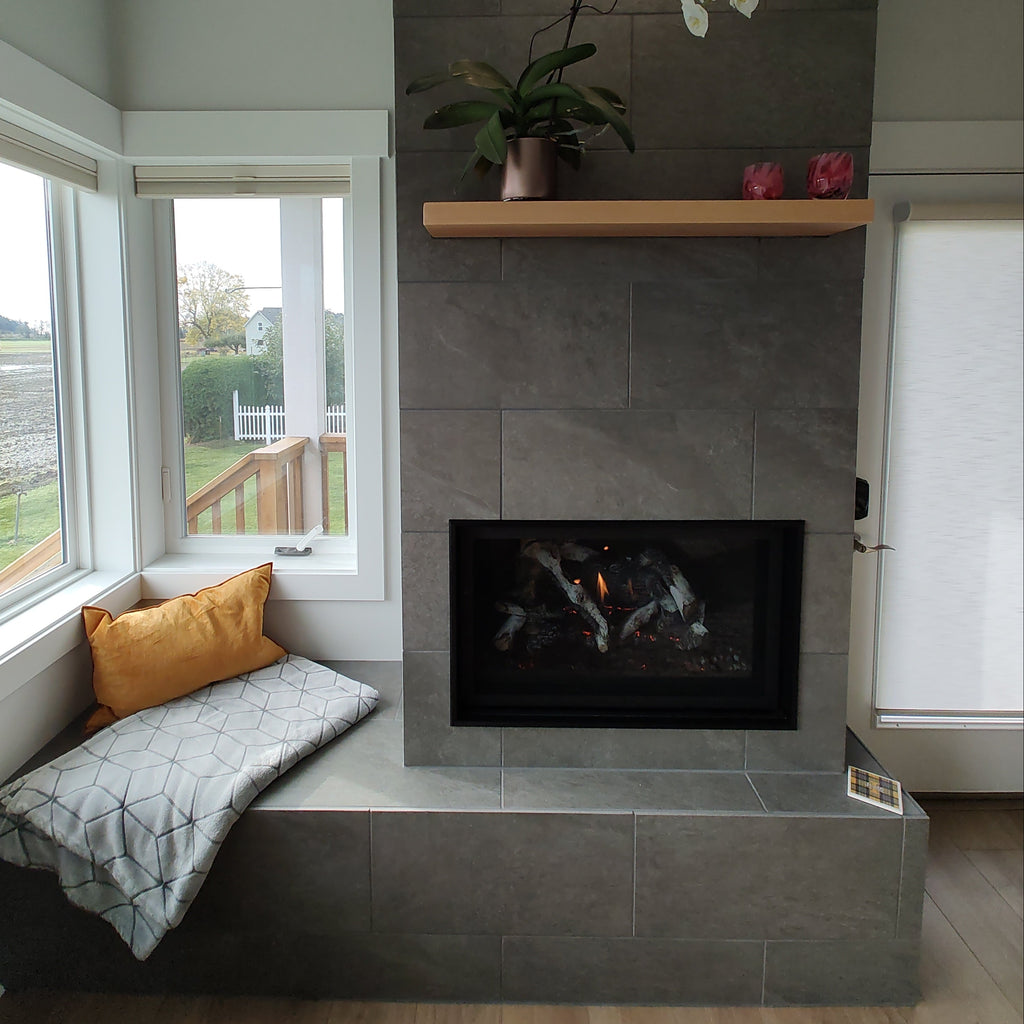 A modern fireplace wall with dark gray stone tiles, a black firebox insert, a light wood mantel with white and pink flowers, and a built-in bench with gray and mustard pillows, shown from an overhead angle.