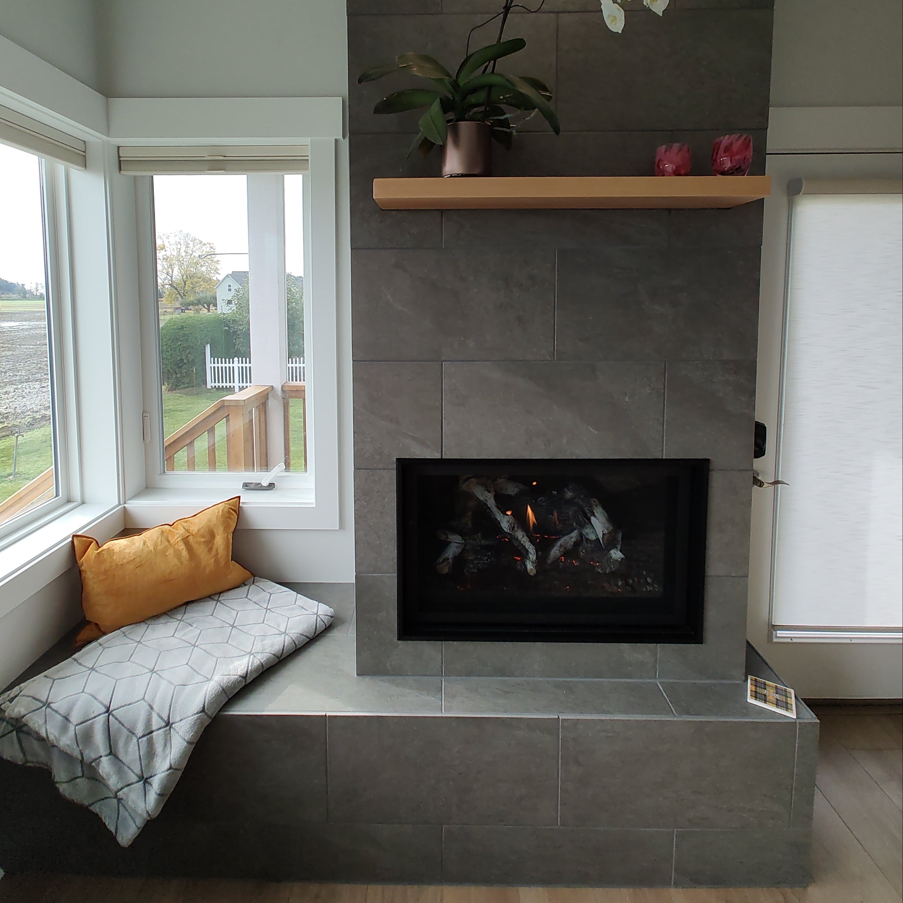 A modern fireplace wall with dark gray stone tiles, a black firebox insert, a light wood mantel with white and pink flowers, and a built-in bench with gray and mustard pillows, shown from an overhead angle.